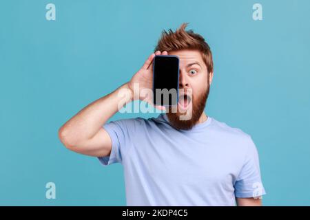 Portrait d'un homme étonné surpris barbu couvrant la moitié du visage avec un téléphone portable et regardant un appareil photo à bouche ouverte, visage choqué. Studio d'intérieur isolé sur fond bleu. Banque D'Images