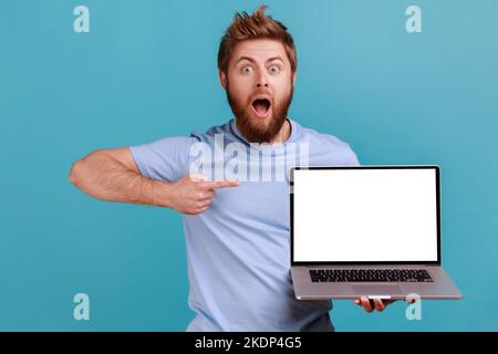 Portrait d'un homme barbu choqué tenant un ordinateur portable et pointant vers un écran vide, surpris par l'application informatique, regardant l'appareil photo. Studio d'intérieur isolé sur fond bleu. Banque D'Images