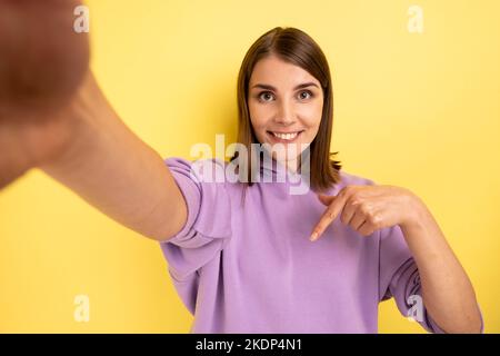 Souriante femme attrayante prenant selfie, point de vue de la photo, pointant le doigt vers le bas, recommander de souscrire son vlog, portant le pull à capuche violet. Studio d'intérieur isolé sur fond jaune. Banque D'Images