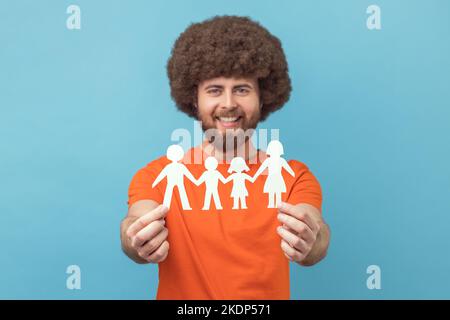 Portrait d'homme souriant satisfait avec la coiffure afro portant un T-shirt orange tenant des symboles de papier de la famille, regardant l'appareil photo, la relation. Studio d'intérieur isolé sur fond bleu. Banque D'Images