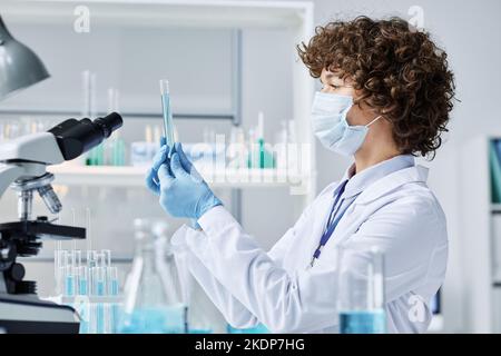 Jeune virologiste dans un masque de protection et des gants tenant un flacon avec une substance liquide bleue tout en l'étudiant en laboratoire clinique Banque D'Images