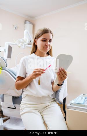 Une belle femme dans un T-shirt blanc est assise dans un cabinet dentaire, regarde dans le miroir, sourit et tient une brosse à dents dans ses mains. Fille avec bretelles montre Banque D'Images