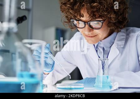 Jeune scientifique ou chimiste féminine en blouse de laboratoire, gants et lunettes qui dépose la substance liquide dans la boîte de Pétri sur le lieu de travail Banque D'Images