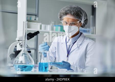 Jeune chercheuse en vêtements de travail de protection faisant des expériences cliniques tout en mélangeant deux substances liquides dans la boîte de Petri Banque D'Images