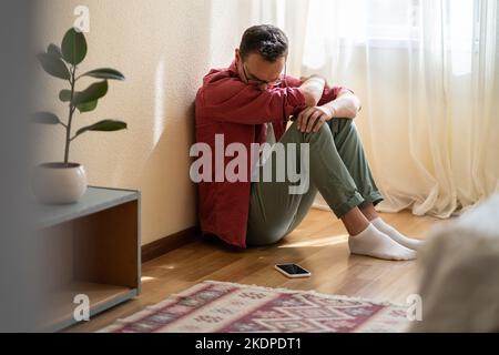 Un homme caucasien déprimé est assis sur le plancher de l'appartement et regarde malheureusement le téléphone mobile en attente d'appel Banque D'Images