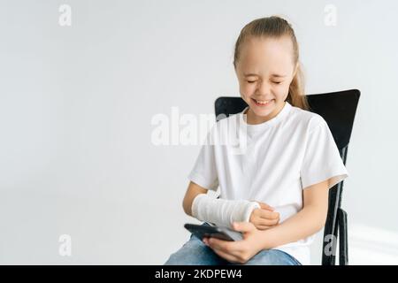 Portrait de la petite fille blonde en riant avec bras cassé enveloppé dans un bandage en plâtre blanc en utilisant le téléphone portable assis à la chaise sur fond blanc. Banque D'Images