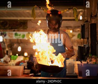 Prenez-le pendant qu'il fait chaud. Un vendeur d'aliments non identifiable portant un casque tout en flamant quelque chose dans une casserole. Banque D'Images