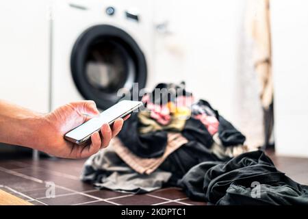 Machine à laver, téléphone et buanderie. Technologie Smart Home et smartphone. Personne utilisant un téléphone cellulaire ou un SMS pendant que vous faites des tâches domestiques. Banque D'Images