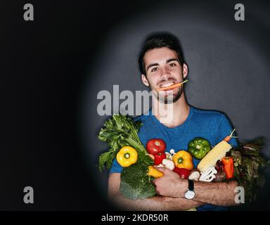 Sain à l'extérieur commence à l'intérieur. Portrait en studio d'un jeune homme portant une quantité de légumes sains sur un fond sombre. Banque D'Images