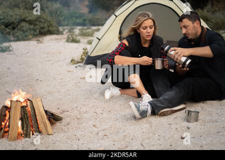 Deux voyageurs boivent du café. Pique-nique sur la plage. Homme versant du thé à partir de thermos. Femme tenant une tasse en métal avec une boisson vapeur. Moment de vie à Banque D'Images