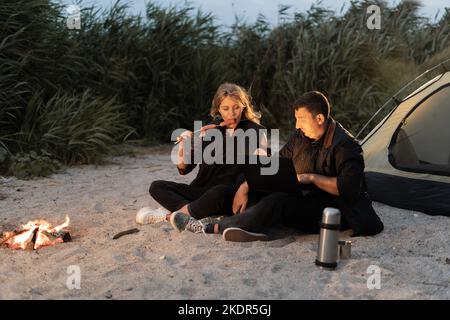 Un couple utilisant un ordinateur à l'extérieur d'une tente lors d'un voyage de camping sur la plage le soir en été. Détente à la tente de camping près de la mer au coucher du soleil. freelance Banque D'Images