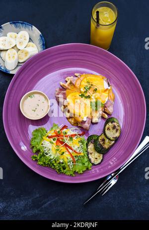 Œufs Benedict (œufs pochés) sur jambon grillé avec sauce hollande sur gaufres belges avec courgettes grillées avec assiette de banane et une tasse de jus de passion Banque D'Images