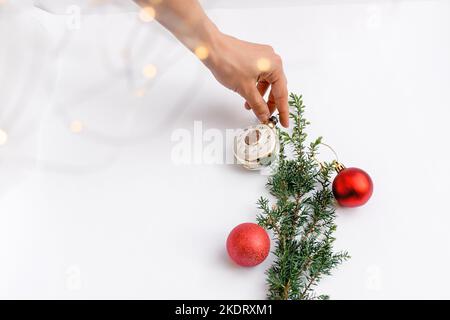 Une femme décorera une branche d'arbre de Noël avec des jouets ronds rouges et une horloge de Noël sur fond blanc. Bokeh jaune brillant. Espace de copie pour le texte et les cartes de vœux. Mise au point sélective sur le premier plan. Banque D'Images