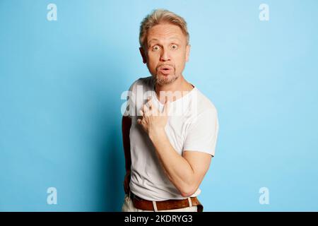 Portrait d'un homme mature en T-shirt blanc posé isolé sur fond bleu de studio. Visage choqué Banque D'Images