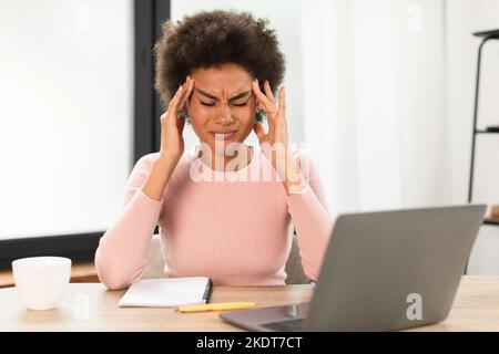 Triste femme afro-américaine millénaire fatiguée avec un ordinateur portable souffrant de maux de tête, appuie les mains sur les temples Banque D'Images