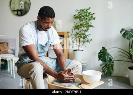 Portrait d'un jeune homme noir utilisant la roue de poterie et la création de céramiques faites à la main dans un studio confortable avec des plantes vertes Banque D'Images