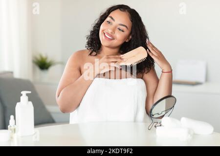 Bonne femme africaine américaine plus taille se brossant les cheveux après la douche, assise enveloppée dans une serviette, tenant une brosse en bois Banque D'Images