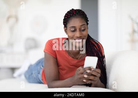 Communication mobile. Message de femme noire souriante sur le smartphone en étant allongé sur le canapé Banque D'Images