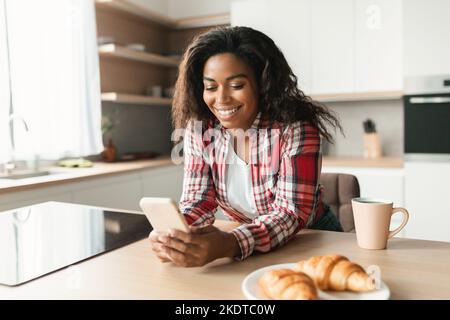 Jeune femme noire gaie dans une dactylographie décontractée sur un smartphone, bavarder, prendre le petit déjeuner, bonne matinée Banque D'Images