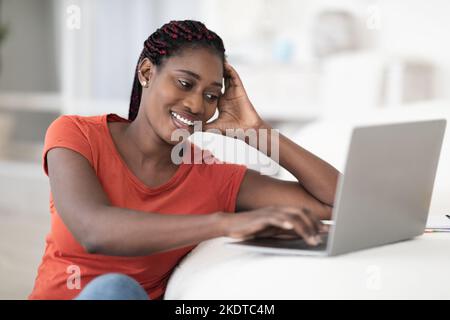 Joyeuse jeune femme noire assise au bureau et utilisant un ordinateur portable à la maison Banque D'Images