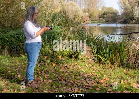 Femme adulte debout vérifiant la caméra avant de prendre une photo, lac entouré d'arbres et de végétation sauvage en arrière-plan, réserve naturelle Eijsden Beemden Banque D'Images