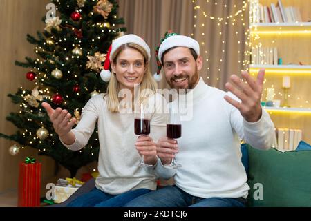 Portrait du nouvel an festif d'un jeune couple. Ils sont assis sur le canapé près de l'arbre de Noël dans des chapeaux rouges du Père Noël, tenant des verres de vin dans leurs mains, invitant les invités avec des gestes de main. Banque D'Images