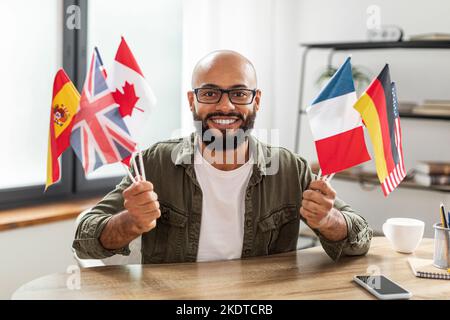 Heureux latin mature étudiant à l'école de langue en ligne, assis au bureau avec des drapeaux de différents pays Banque D'Images