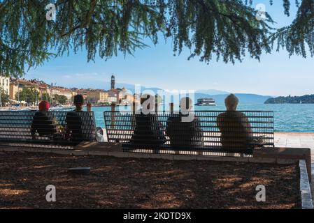 Les personnes âgées l'été, vue arrière d'un groupe de personnes âgées assis sur un banc et regardant la ville pittoresque du lac de Garde de Salo, Lombardie, Italie Banque D'Images