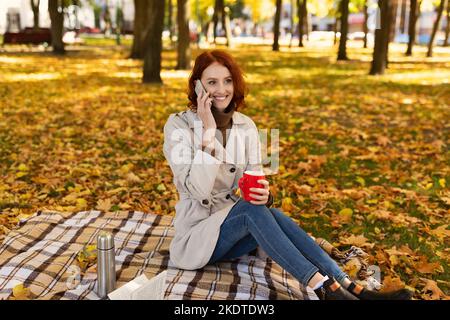 Bonne femme du millénaire caucasien avec cheveux rouges en imperméable assis sur le tissu écossais avec thermos, tient la tasse avec la boisson chaude Banque D'Images