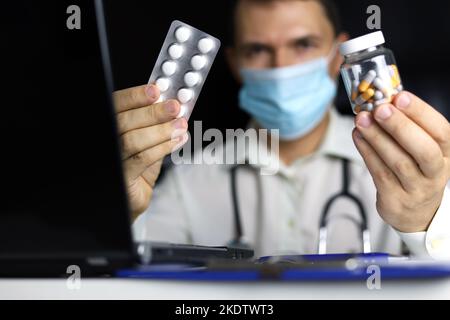 Pilules et capsules à la main du médecin dans le masque médical assis à l'ordinateur portable dans le bureau. Homme remplissant l'histoire médicale, concept de prescription Banque D'Images