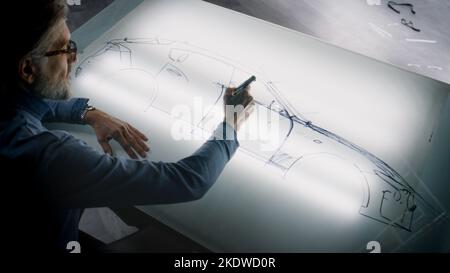 Un homme de conception automobile senior dessinant un plan avec un marqueur à la table lumineuse, développant un design futuriste de voiture électrique. Il travaille dans un studio de développement de conception automobile moderne. Banque D'Images