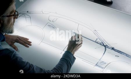 Un homme de conception automobile senior dessinant un plan avec un marqueur à la table lumineuse, développant un design futuriste de voiture électrique. Il travaille dans un studio de développement de conception automobile moderne. Banque D'Images