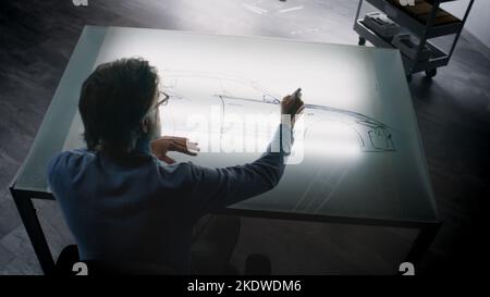 Un homme de conception automobile senior dessinant un plan avec un marqueur à la table lumineuse, développant un design futuriste de voiture électrique. Il travaille dans un studio de développement de conception automobile moderne. Banque D'Images