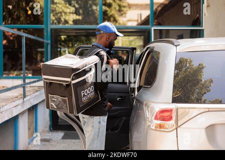 Jeune homme livrant une commande de nourriture de bureau en voiture. Employé du service de livraison de repas à emporter tenant un sac à dos thermique avec un repas, debout près du véhicule à l'extérieur, vue latérale vue moyenne Banque D'Images