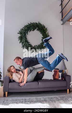 Amoureux, fous couple d'amoureux de s'amuser à la couronne de Noël. Femme élégante avec rouge à lèvres repose sur un canapé, tient le cadeau dans les mains, homme millénaire jum Banque D'Images