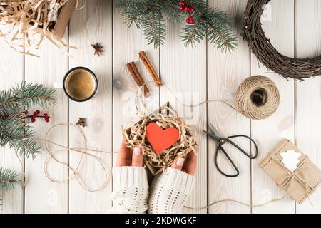 Femme coupée mains dans chandail blanc tenant une boîte en carton ouverte avec coeur rouge dans la décoration de Noël de cannelle, ciseaux, cadeaux et branches de sapin Banque D'Images