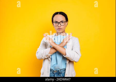 Non, arrêtez, assez. Femme d'affaires sérieuse, strictement hispanique ou brésilienne, portant des lunettes, montrant un geste d'arrêt avec les paumes croisées, regardant sévèrement l'appareil photo, se tenant sur un arrière-plan orange isolé Banque D'Images