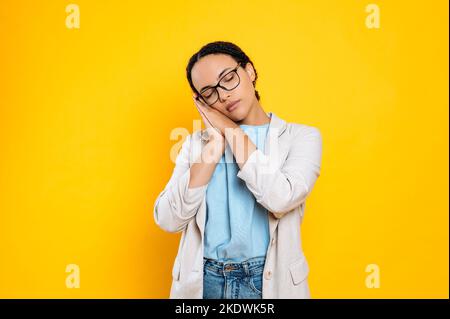 Femme d'affaires fatiguée et surtraitée, portant des lunettes et une veste, montre un geste de sommeil, veut dormir, met sa tête dans ses paumes, ferme ses yeux, se tient sur un fond orange isolé Banque D'Images