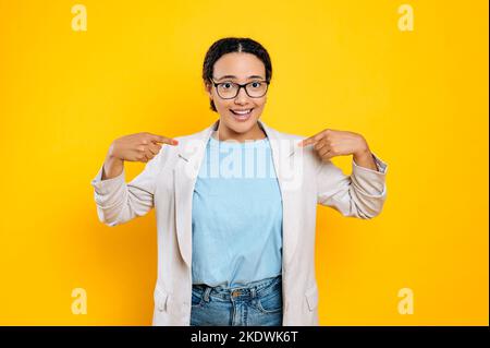 Gai satisfait jeune latino ou hispanique jeune femme dans une veste, femme d'affaires, se montre avec des index, regarde la caméra dans la surprise, sourires, se tient sur un arrière-plan isolé orange Banque D'Images