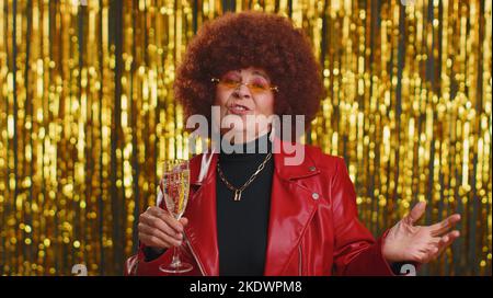 Drôle de femme en blouson rouge écoutant de la musique, souriant dansant à la musique disco de fête, boire du champagne, s'amuser. Grand-mère senior célébrant la victoire, l'anniversaire, Noël isolé sur fond brillant Banque D'Images
