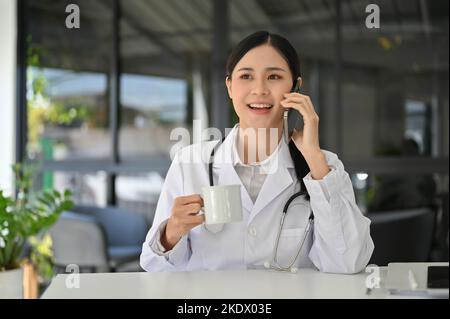 Femme asiatique professionnelle médecin en uniforme discutant avec quelqu'un sur son smartphone et sirotant du café pendant sa pause-café dans le bureau du médecin. Banque D'Images