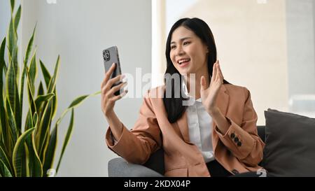 Magnifique jeune femme d'affaires asiatique assise sur un canapé, agitant la main, parlant sur un appel vidéo via son smartphone. Banque D'Images