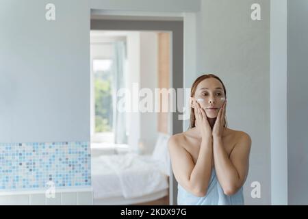 Une jeune femme se tient au miroir dans la salle de bains après une douche et applique de la crème sur son visage. La jeune femme utilise une crème pour le visage. Mise au point douce. Banque D'Images