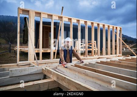 Maison en bois de construction d'architecte mâle. Homme debout sur le chantier dans un casque de sécurité, inspectant la qualité du travail. Banque D'Images