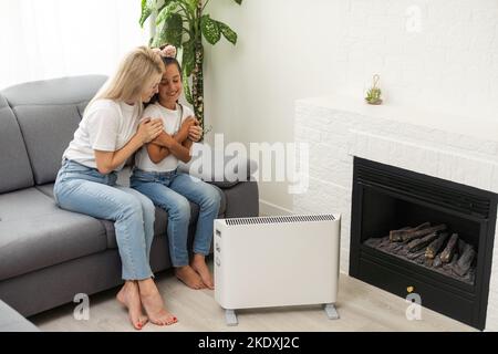 La mère et l'enfant réchauffent les mains près du chauffage électrique à la maison Banque D'Images