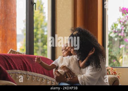 Une mère indienne qui s'embrasse et embrasse une petite fille tout en étant assise sur un canapé confortable dans un salon lumineux à la maison Banque D'Images