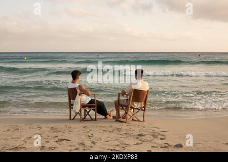 Vue arrière de tout le corps d'amis masculins non reconnaissables en vêtements d'été assis sur des chaises de pêche et en admirant la mer ondulée pittoresque sans fin Banque D'Images