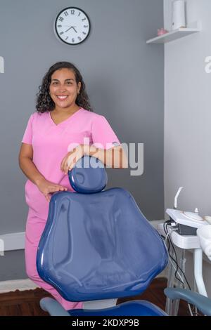 Jeune femme hispanique gaie dentiste avec des cheveux fclés foncés en uniforme rose souriant et regardant la caméra tout en se penchant sur la chaise dentaire dans le clin moderne Banque D'Images