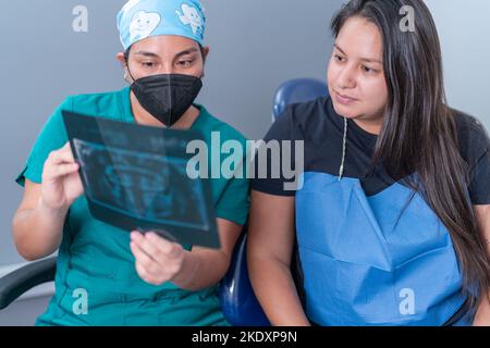 Jeune femme hispanique concentrée dentiste en uniforme et masque montrant l'image de rayons X au patient et expliquant le traitement dans la clinique dentaire moderne Banque D'Images