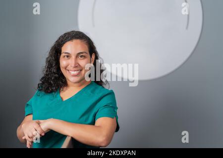Jeune femme hispanique gaie dentiste avec des cheveux fclés foncés en uniforme vert souriant et regardant la caméra tout en se penchant sur la chaise dentaire dans la cli moderne Banque D'Images